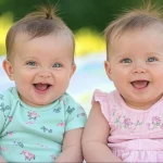Smiling twin babies sitting outdoors on a blanket — one in a mint green onesie and the other in a pastel pink dress, with natural sunlight and greenery in the background.